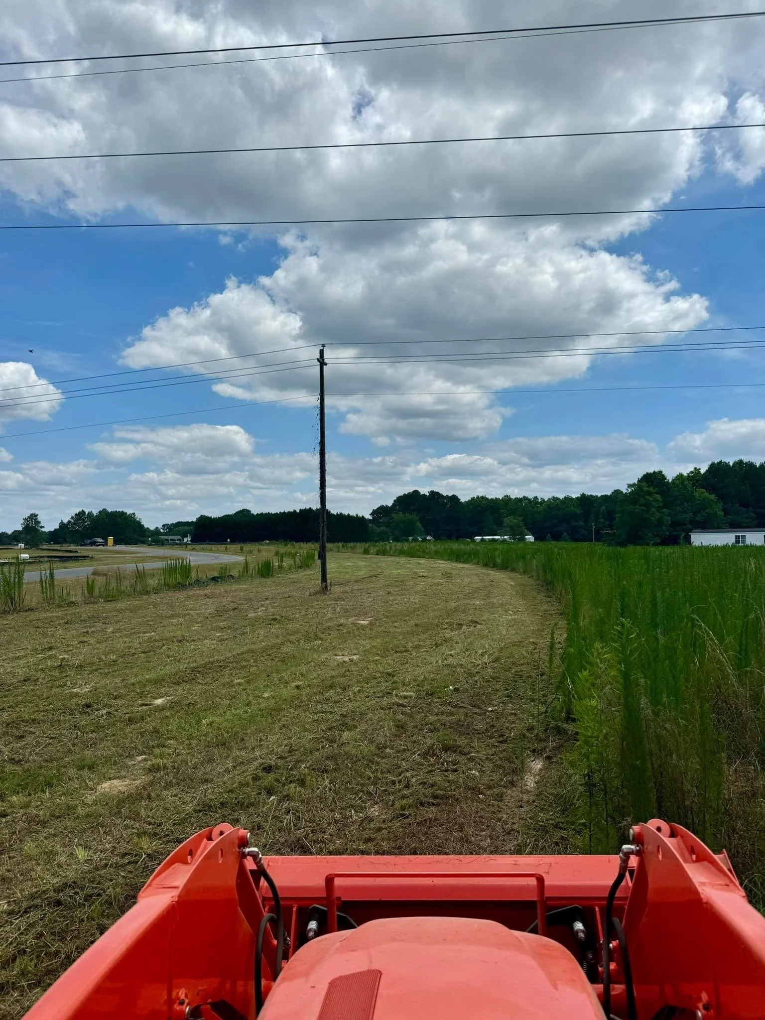 View from the front of a red tractor showing a grassy field, power lines, a partly cloudy sky, and a distant tree line and buildings.
