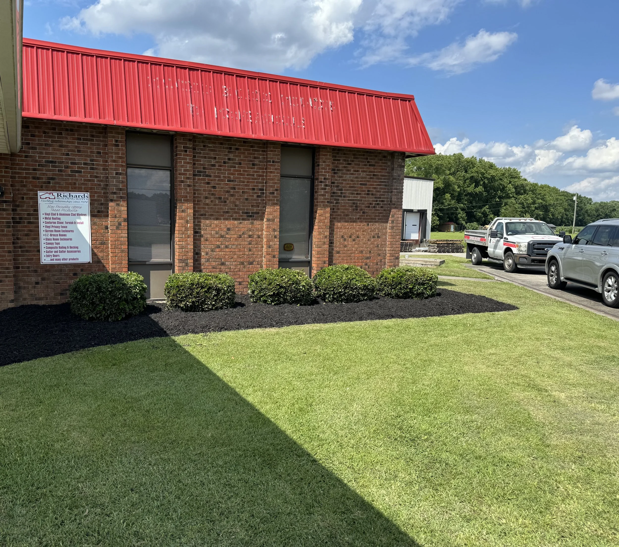Exterior of a brick building with a red metal roof, three small bushes in front, a sign on the left side, and a parking lot with two trucks and one car on the right. Clear blue sky with a few clouds.