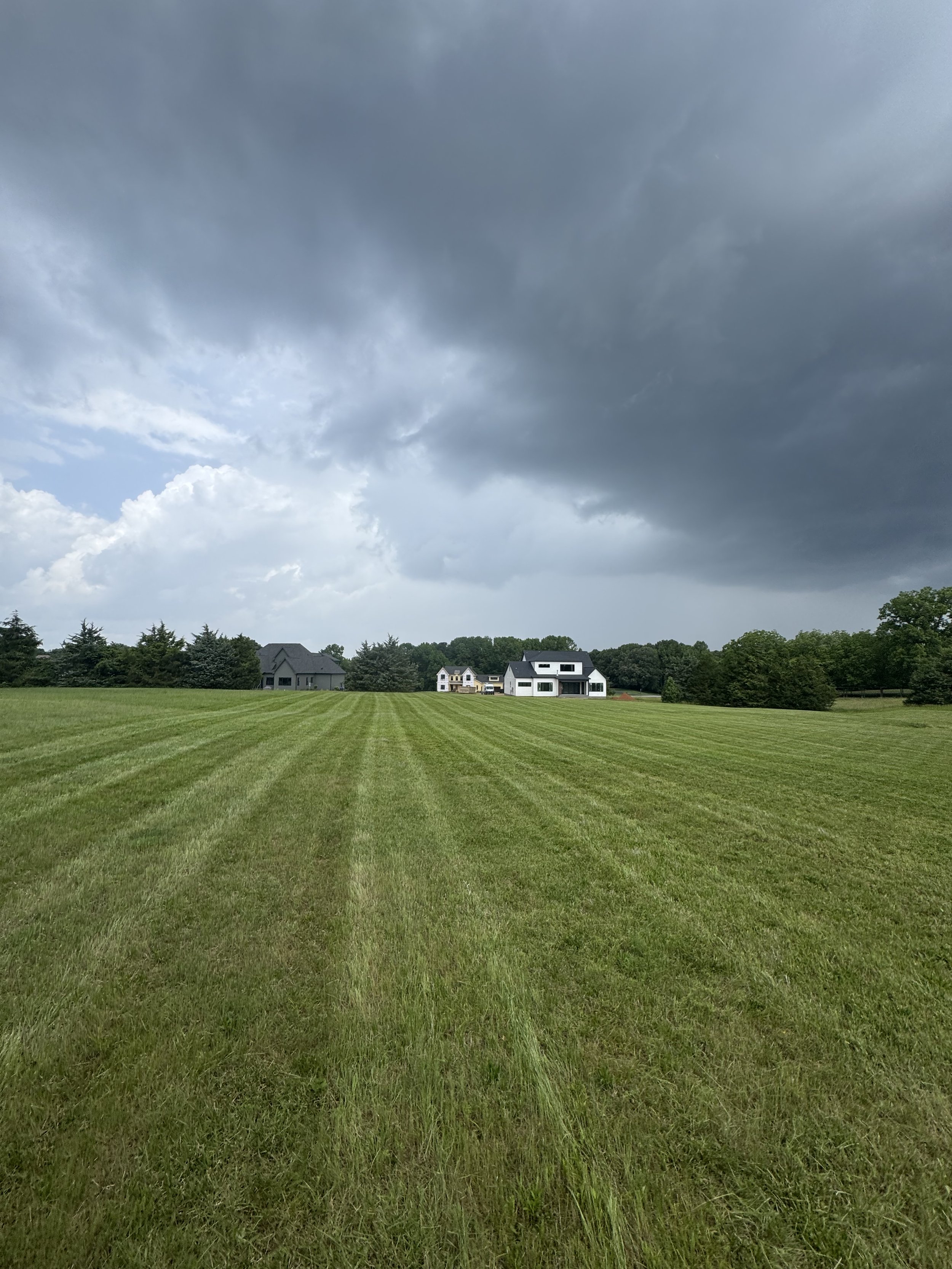 Open green field with striped mowed grass, dark storm clouds in the sky, and houses in the distance at the horizon.