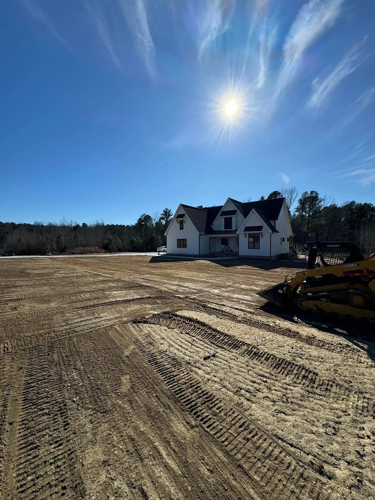 A house under construction with a cleared dirt lot in front and construction equipment nearby, under a bright blue sky with the sun shining.