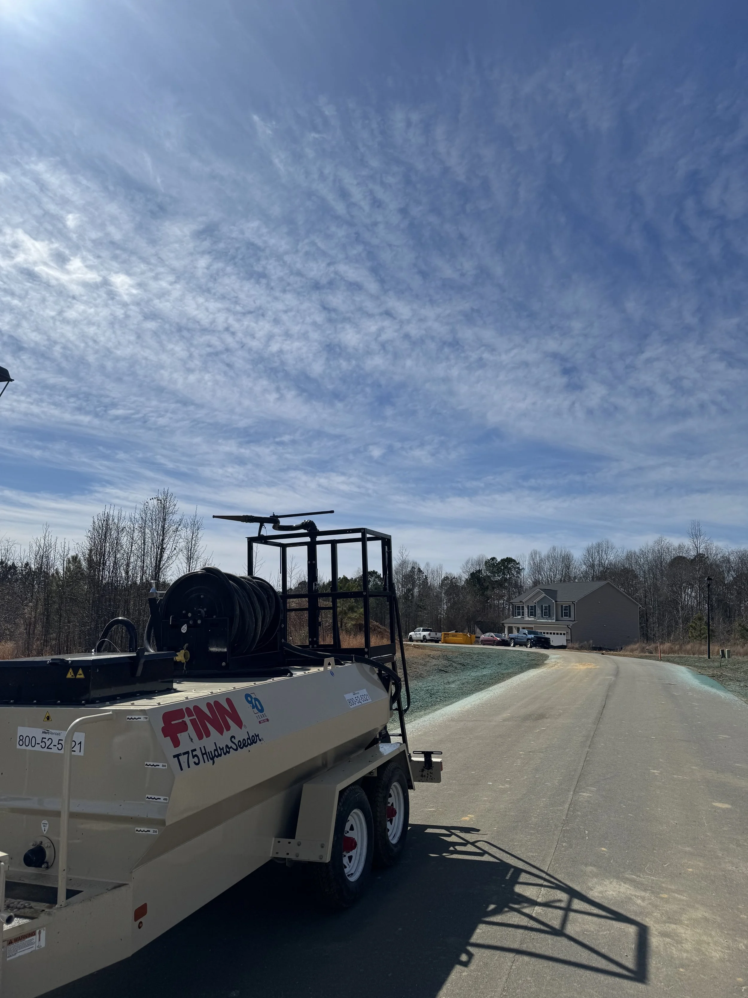 A trailer with firefighting equipment parked on a paved road in a suburban area, with a house and trees in the background, under a blue sky with scattered clouds.