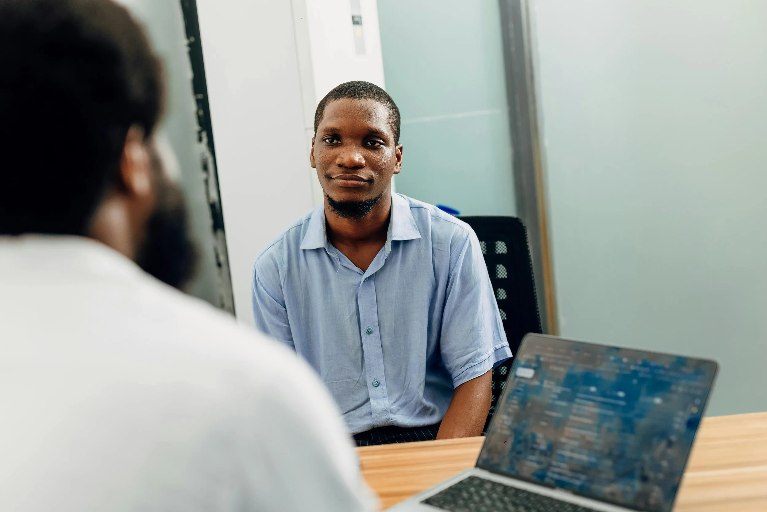 A young man wearing a light blue shirt sits at a table during a conversation in an office setting with a laptop open in front of him.
