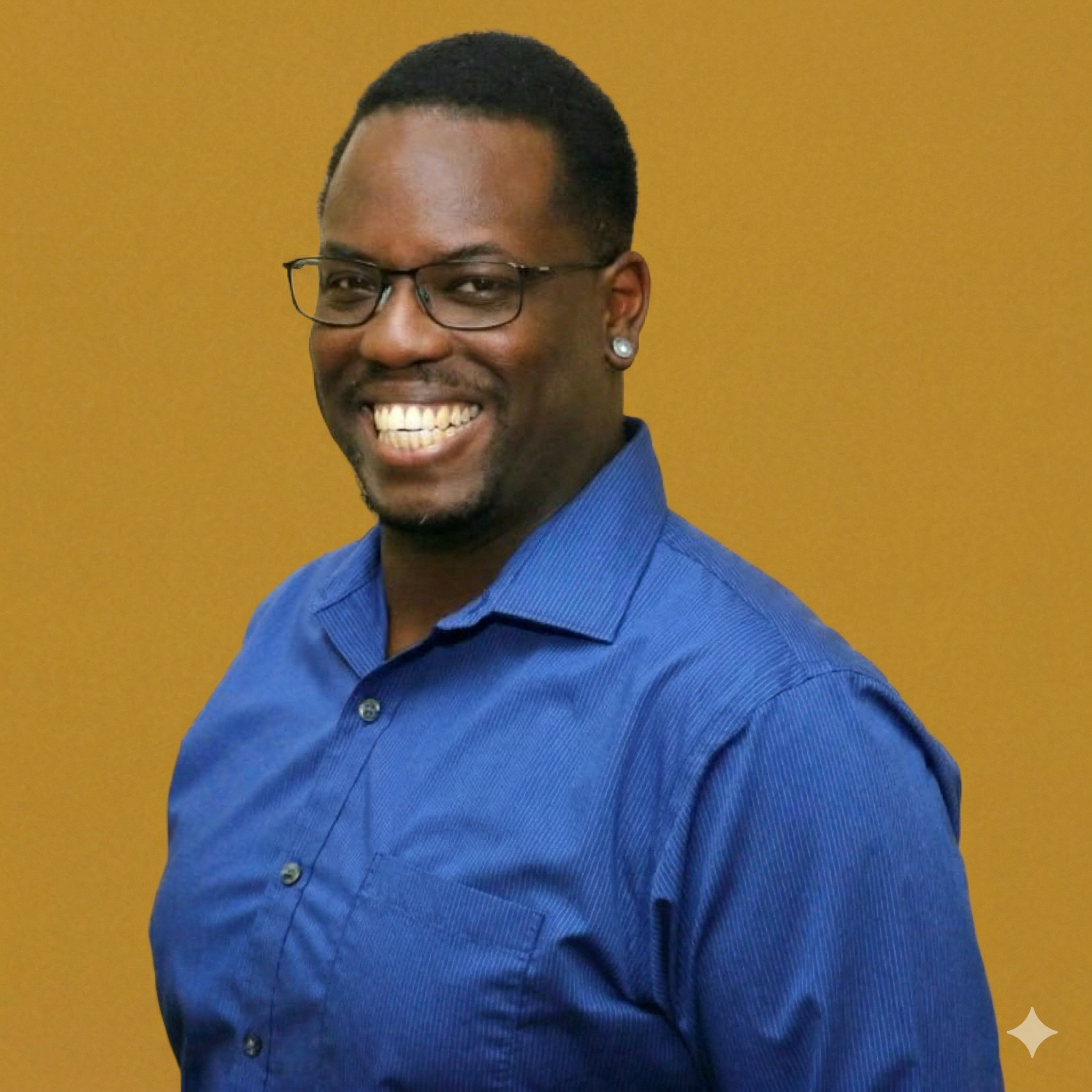 A smiling man with glasses and earrings wearing a blue shirt, posed against a plain yellow background.
