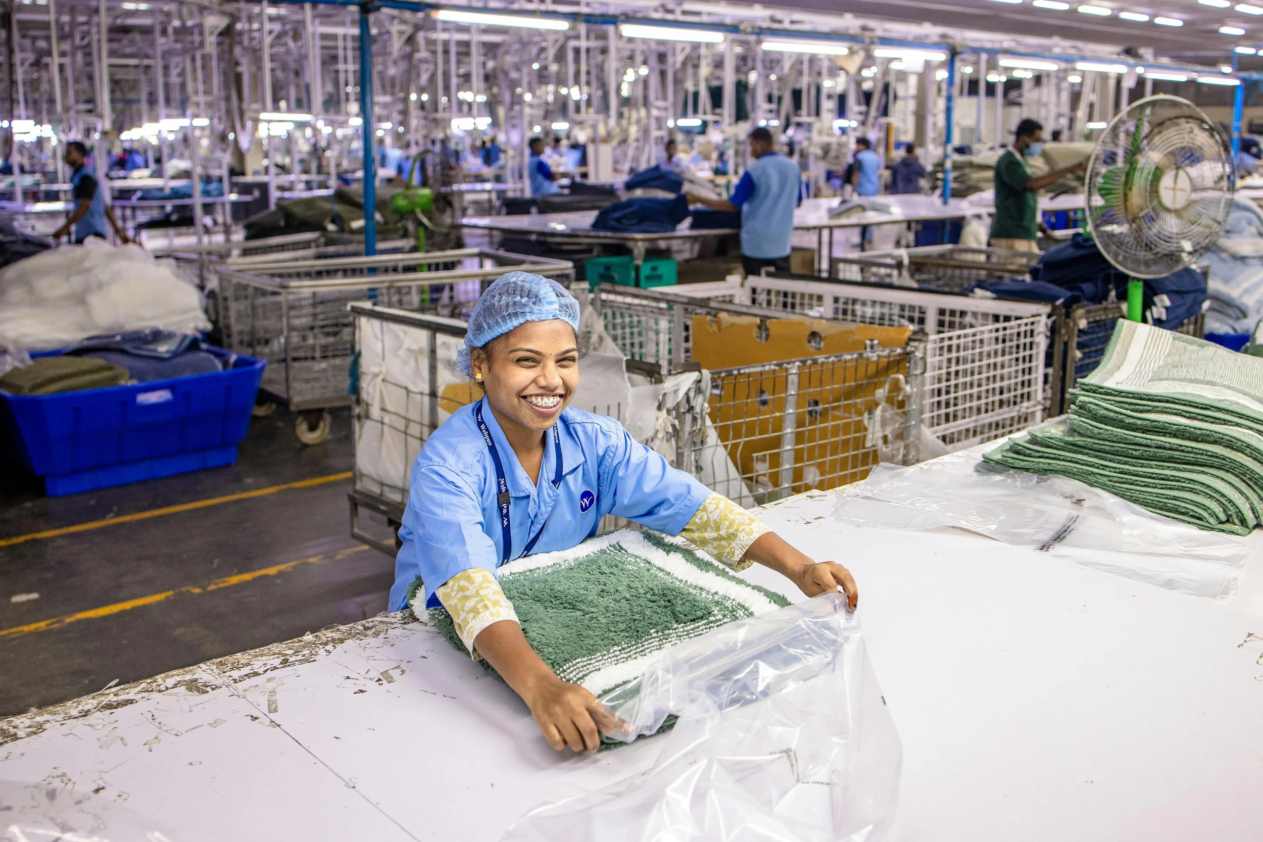 A worker in a laundry factory holding a folded green towel and smiling, with other workers and laundry equipment in the background.