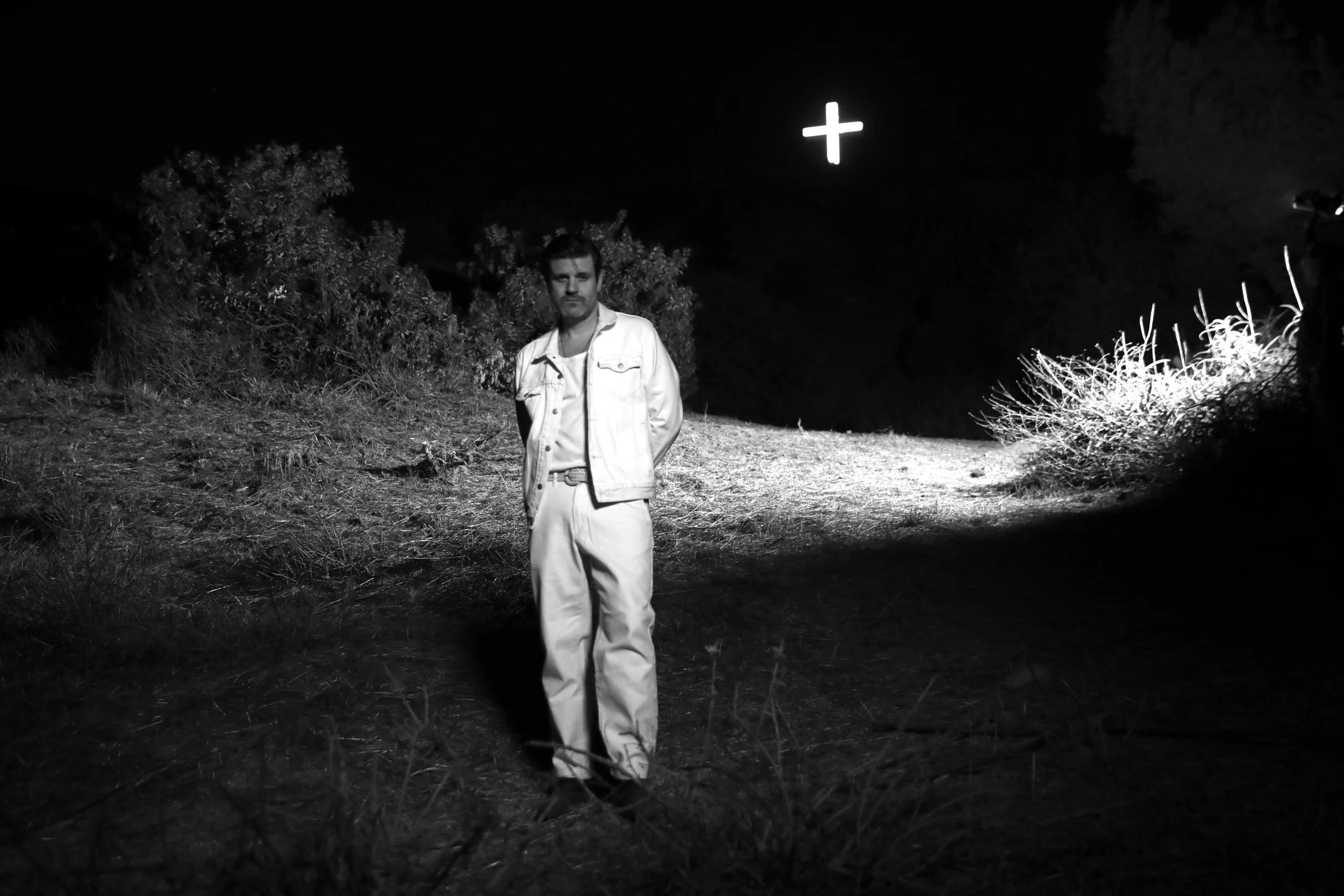 A man standing outdoors at night, illuminated by a flashlight, with bushes and trees around him, and a glowing cross symbol in the dark sky above.
