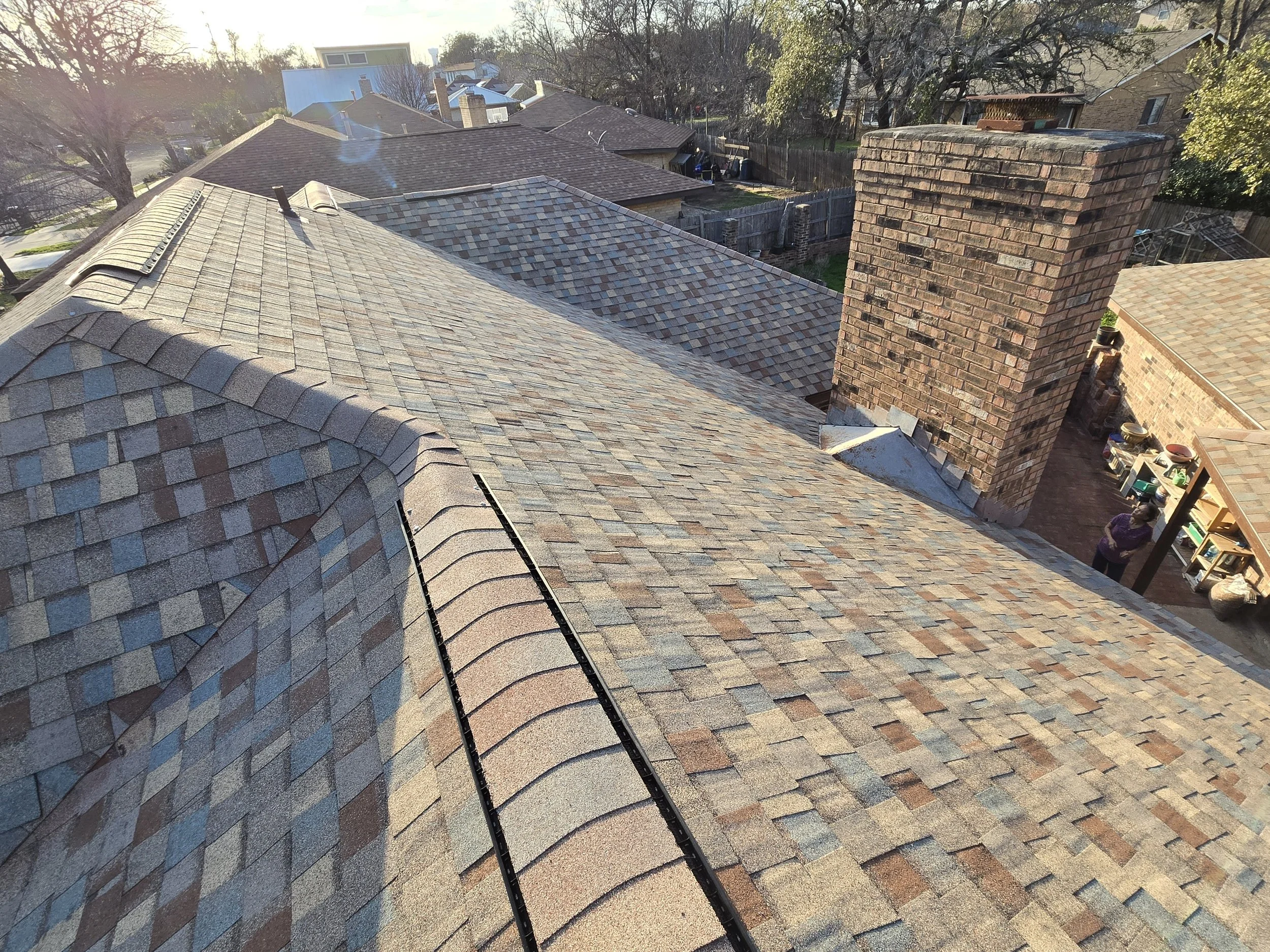 Aerial view of a roof with multicolored asphalt shingles, a brick chimney, and a person standing in a backyard. Sunlight and trees are visible in the distance.