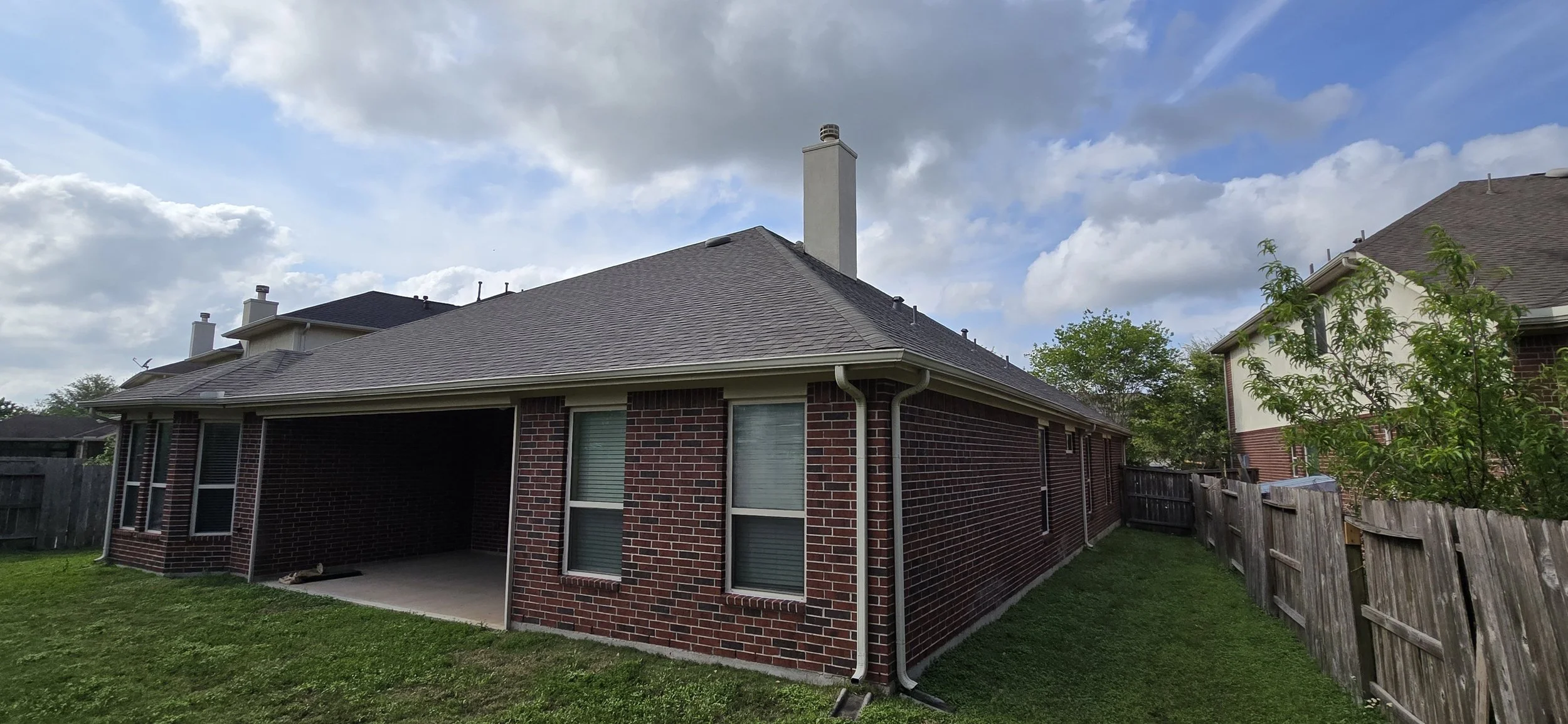 Backyard view of a brick house with a covered patio, gray shingle roof, and a wooden fence, with a lawn and trees under a partly cloudy sky.