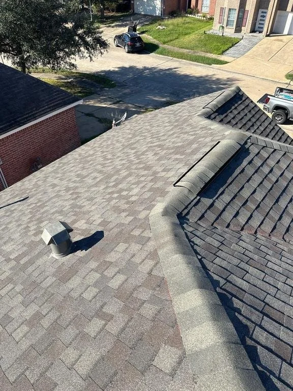 View of a house roof with asphalt shingles, a chimney, and vents, overlooking a driveway with parked cars and neighboring houses.