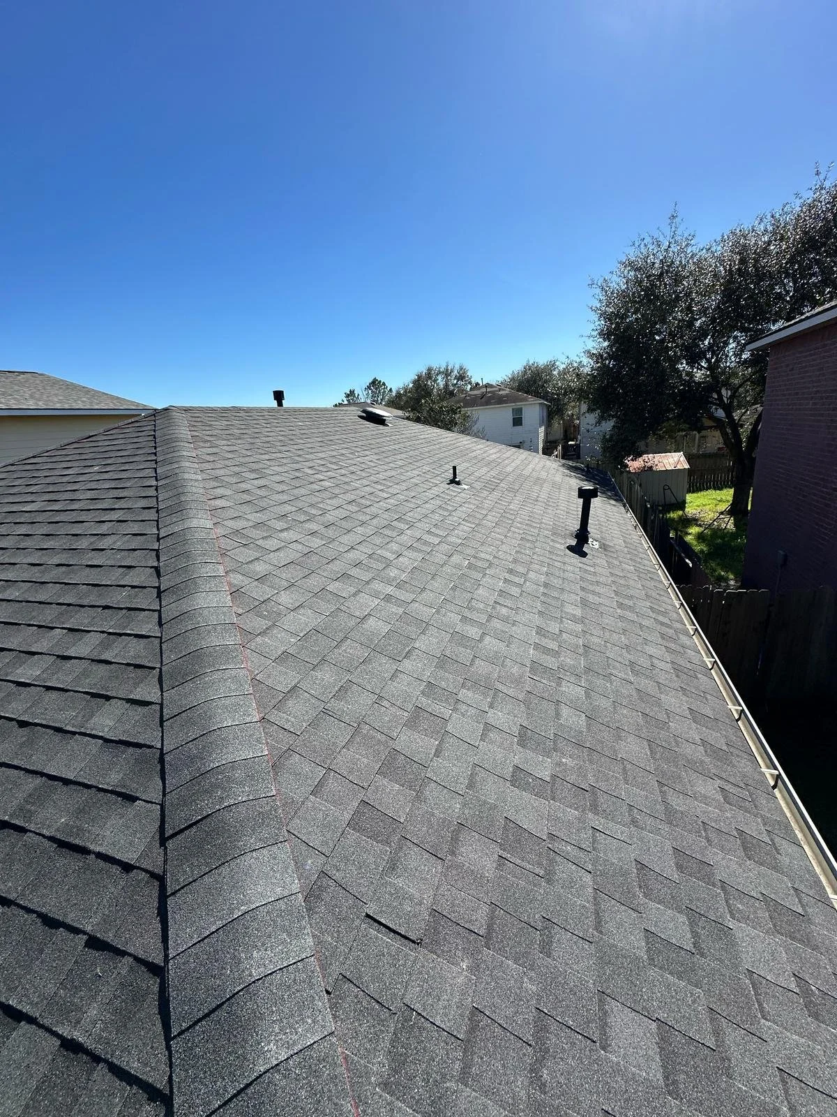 View of a residential rooftop covered with grey asphalt shingles, showing vents and structures, with neighboring houses and trees under a clear blue sky.