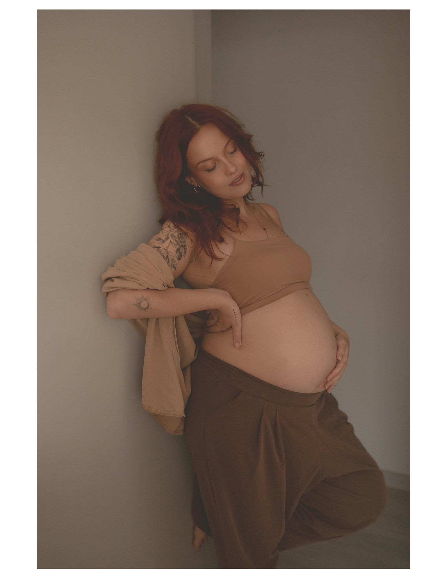 A pregnant woman with red hair, tattoos, wearing a beige top and brown pants, leaning against a plain wall with her eyes closed and hand resting on her belly.