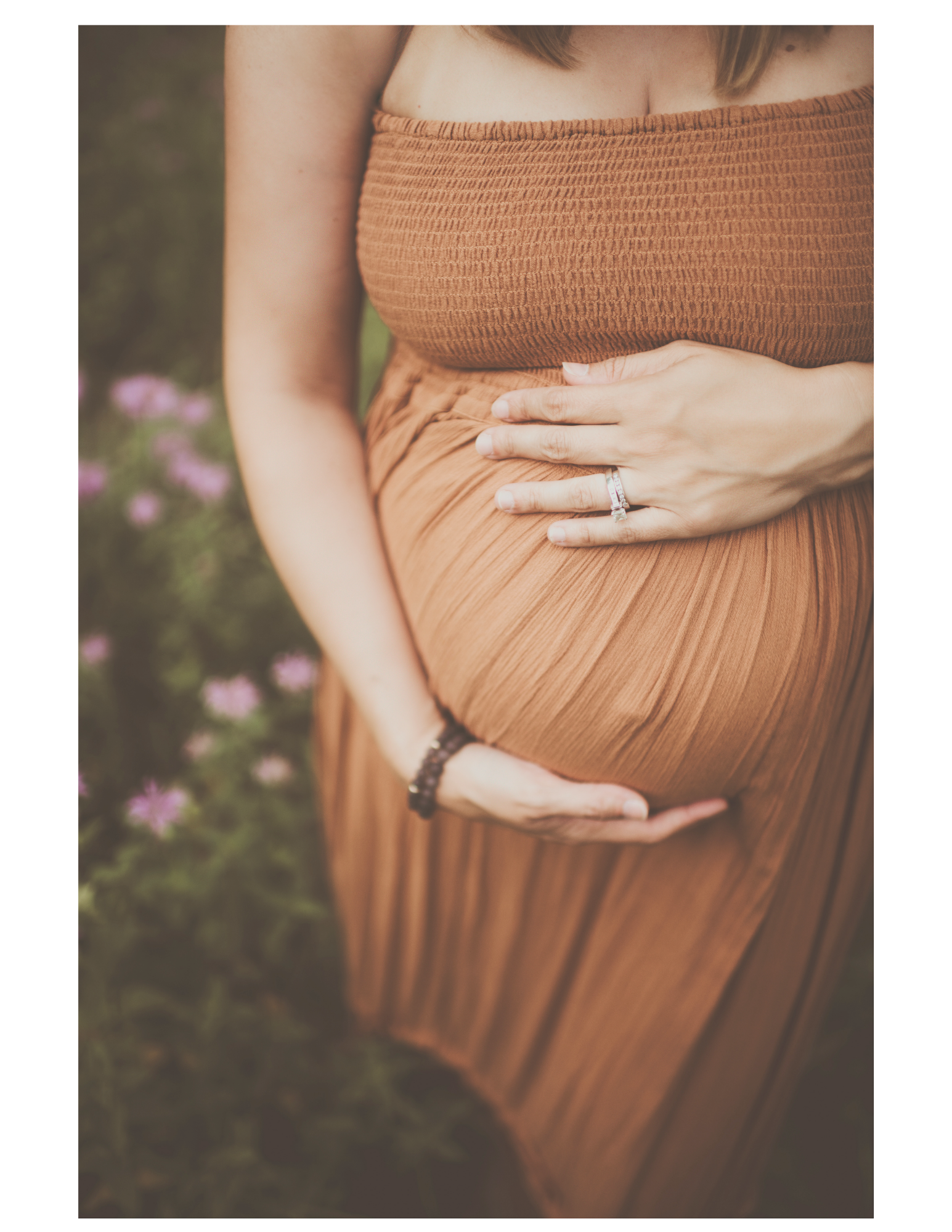 A woman in an earthy orange strapless dress with a textured fabric gently holding her pregnant belly, standing outdoors with blurred pink flowers and grass in the background.