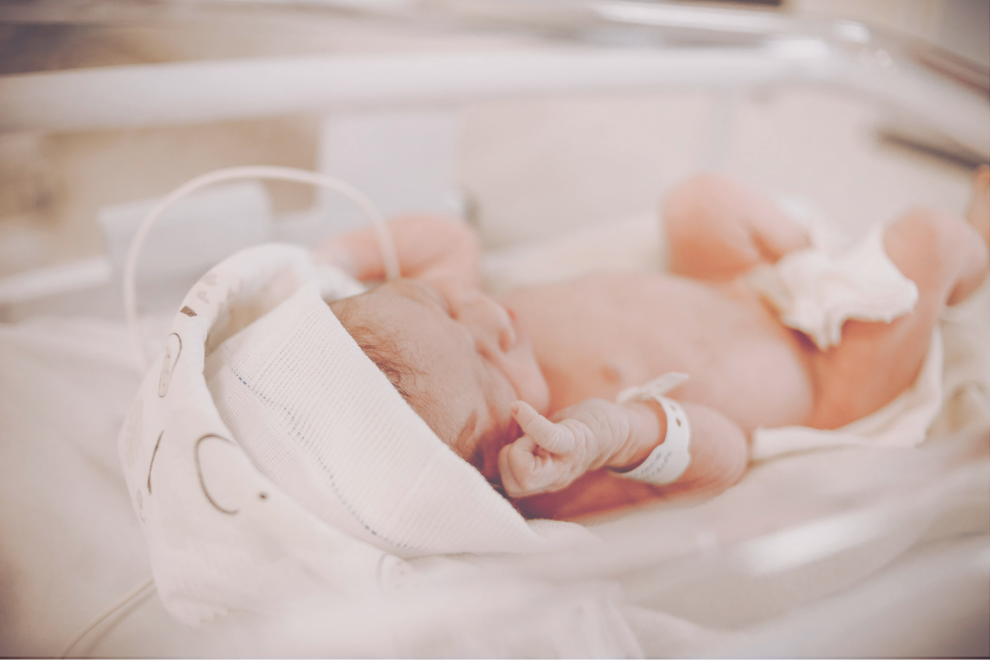 Newborn baby in a hospital setting lying in a crib, wearing a small cap and a hospital bracelet.