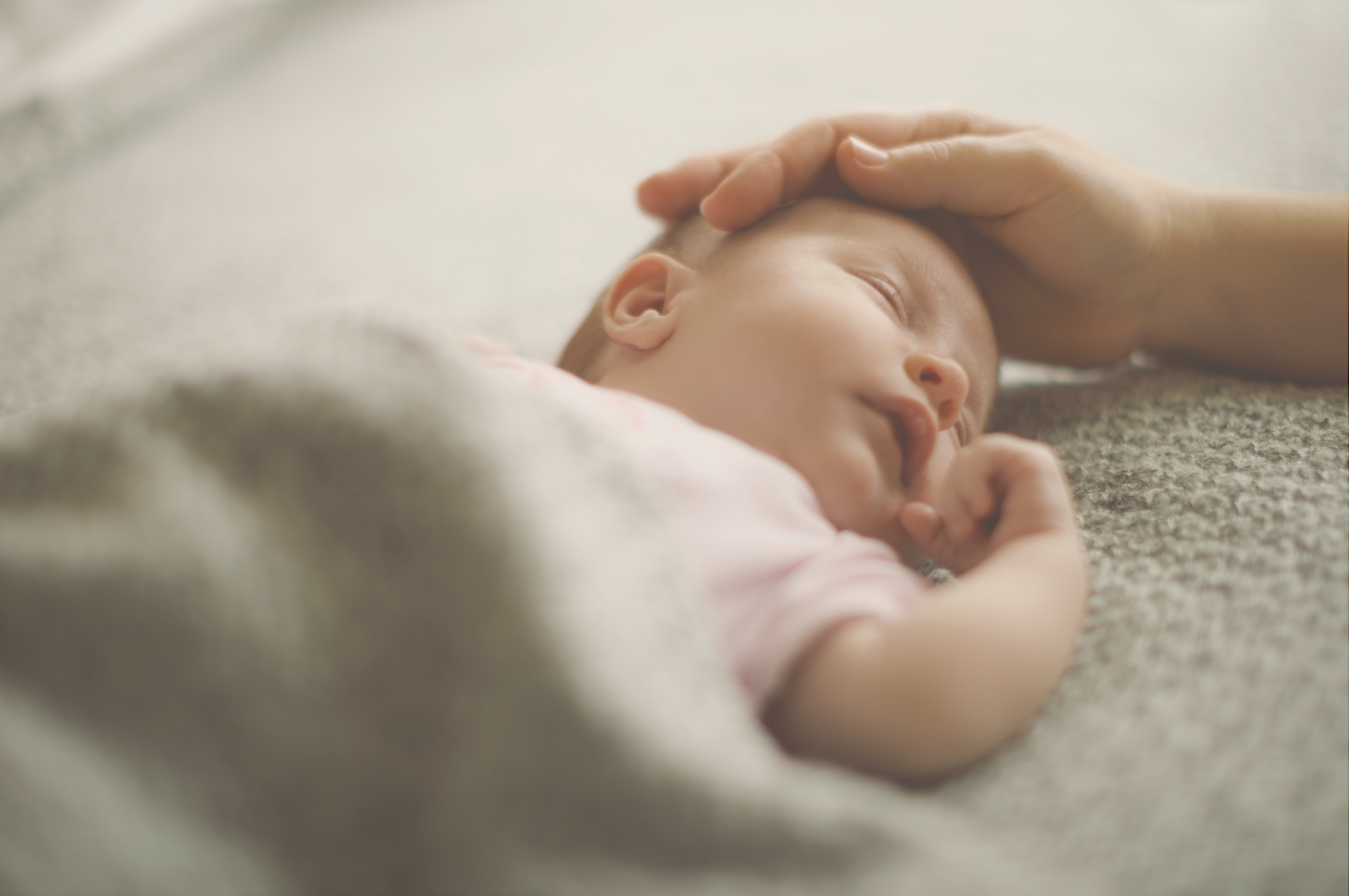 Close-up of a sleeping baby lying on a soft surface, with an adult hand gently resting on their head, showing a moment of tenderness.