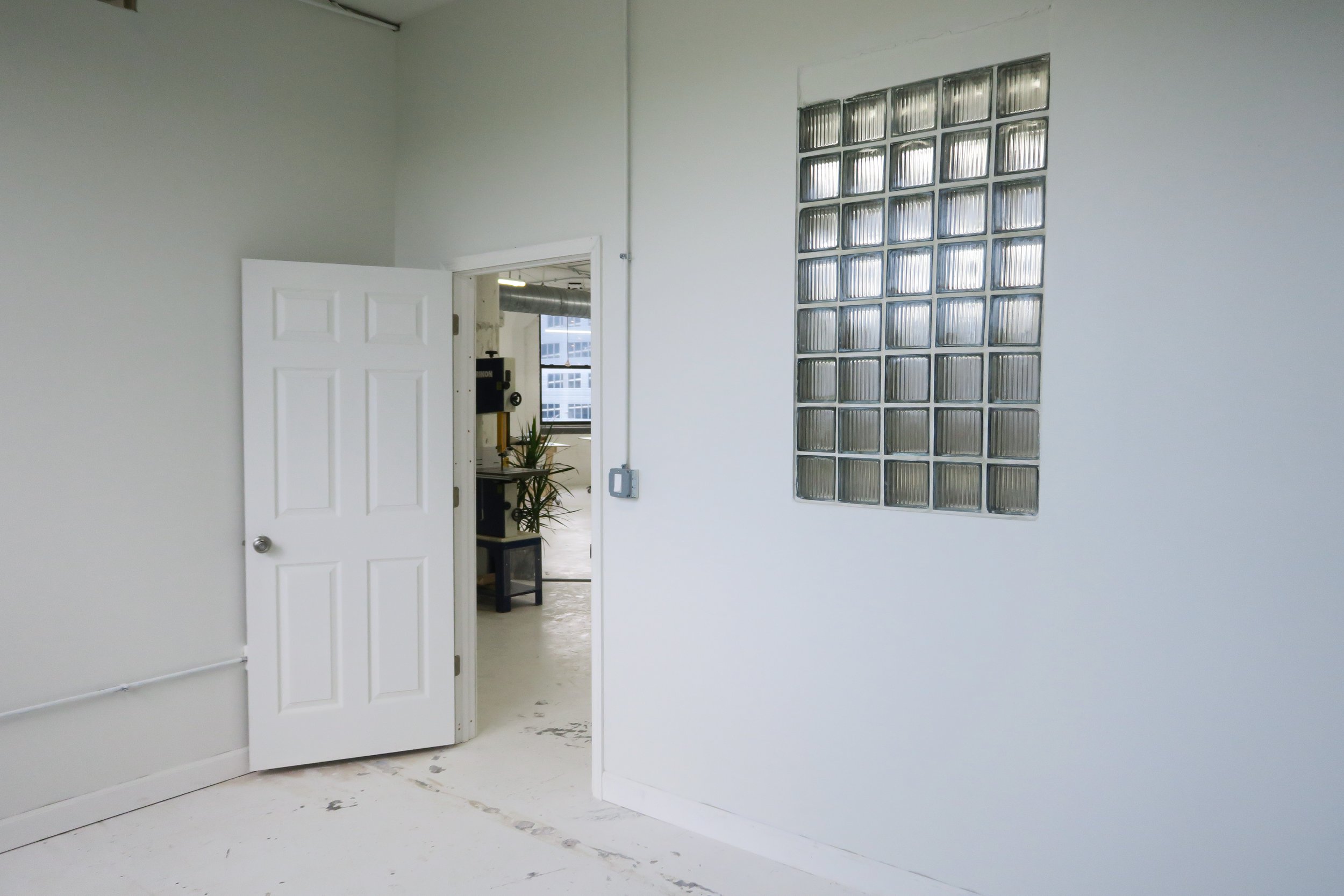 An interior space with white walls and a partially open white door, revealing a room with a plant, a black piece of furniture, and a window. There is a glass block window on the right wall.