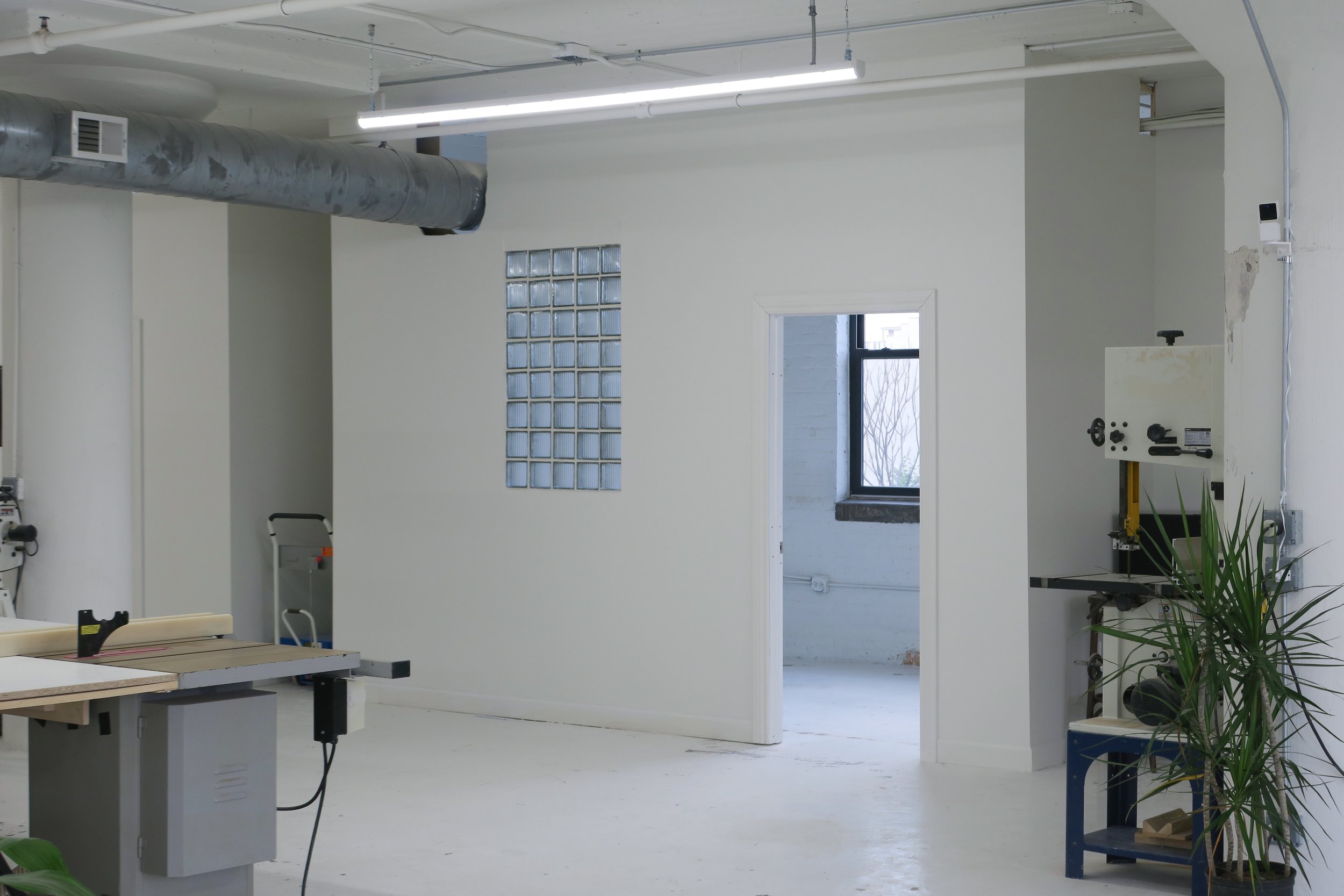 A spacious, industrial-style workshop with white walls, a large window, lawn in the background, fluorescent ceiling lights, and various tools and equipment, including a saw and a drill press, along with a potted plant.