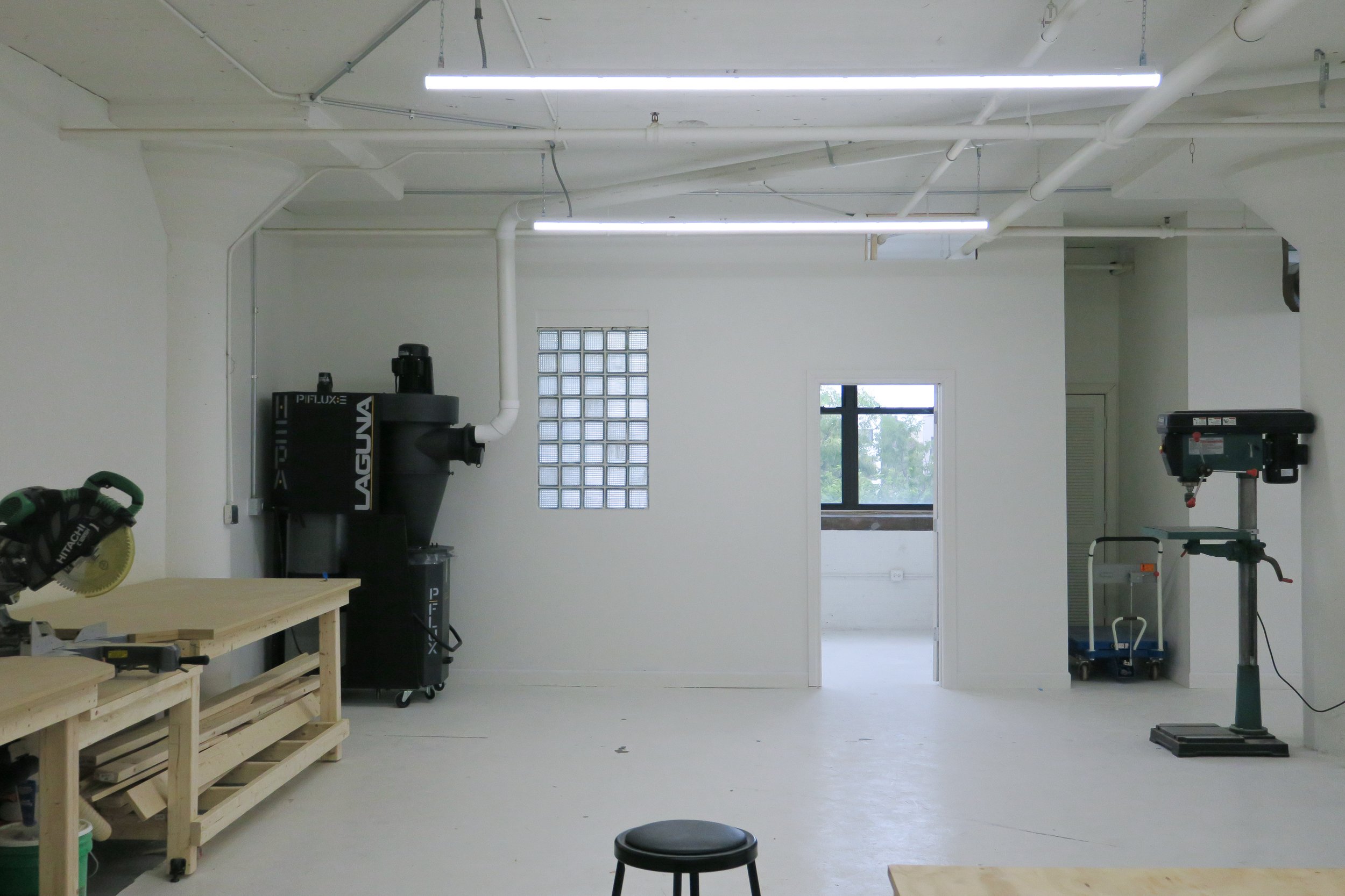 Empty workshop room with workbenches, power tools, a small stool, and natural light coming through windows.