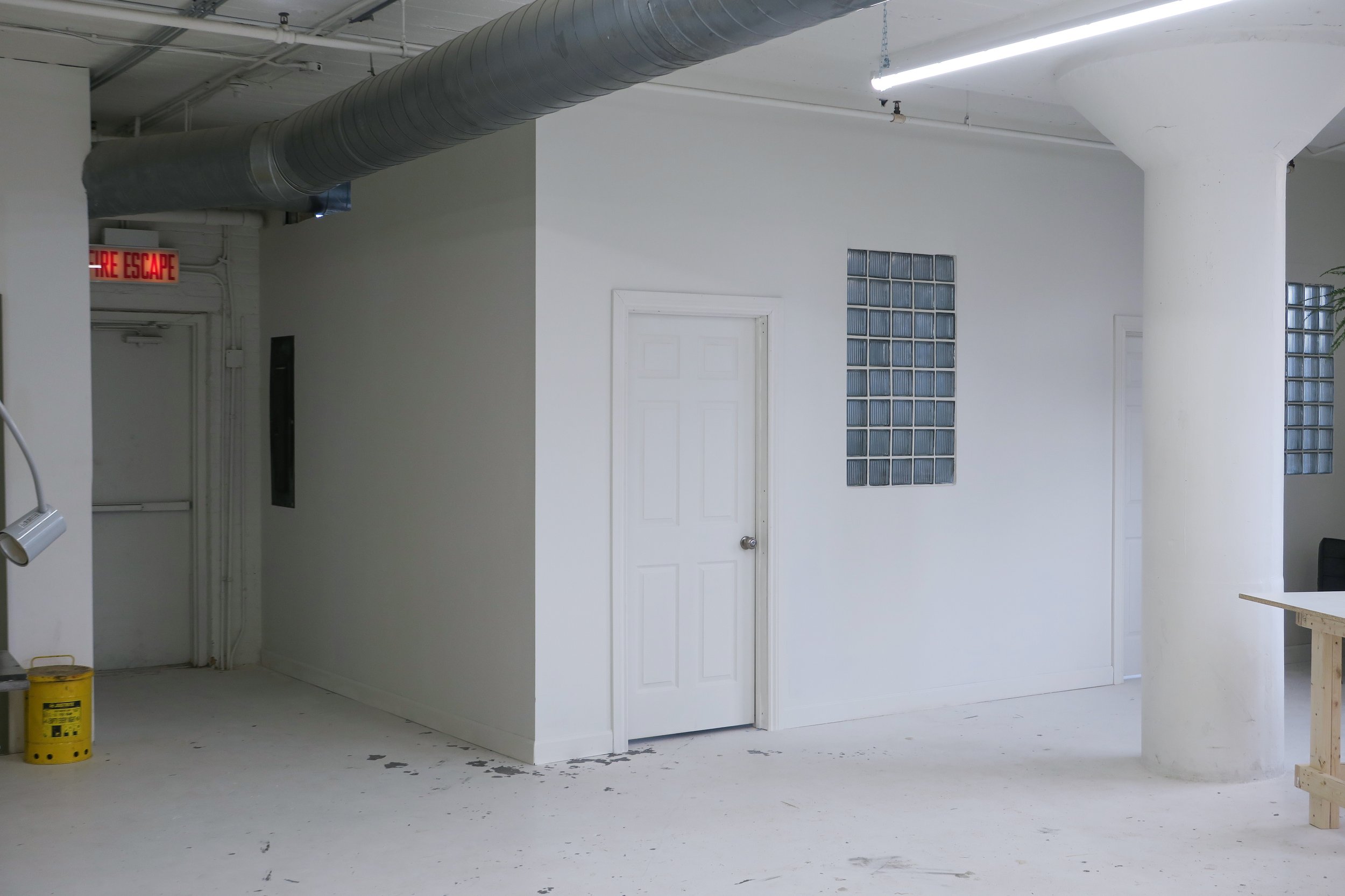 Interior of a mostly white room with a closed door, glass block windows, a large white support column, an exposed duct, fire exit sign, and construction materials.