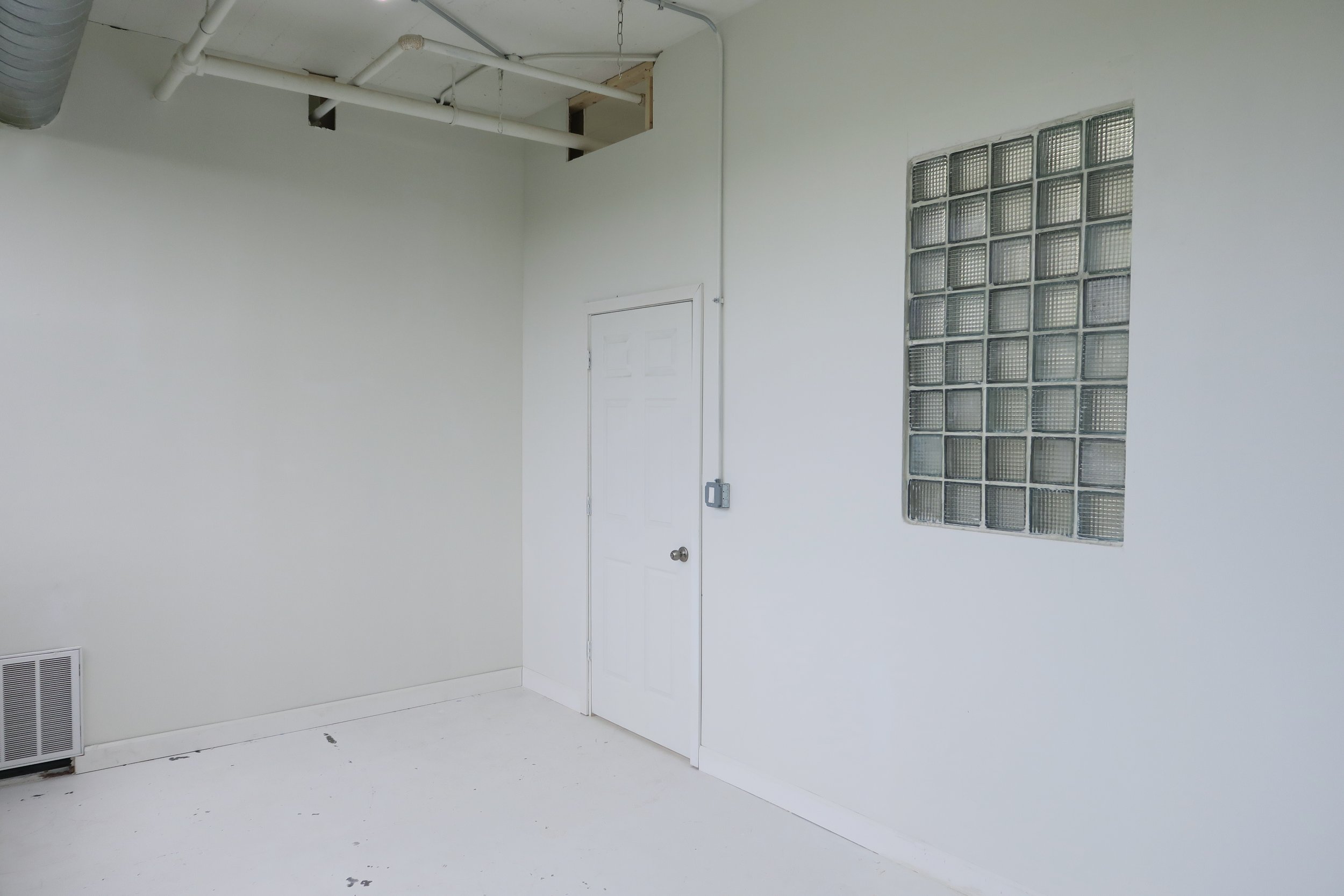 Empty white room with a glass block window, a white door, and an exposed ceiling with white pipes and ductwork.