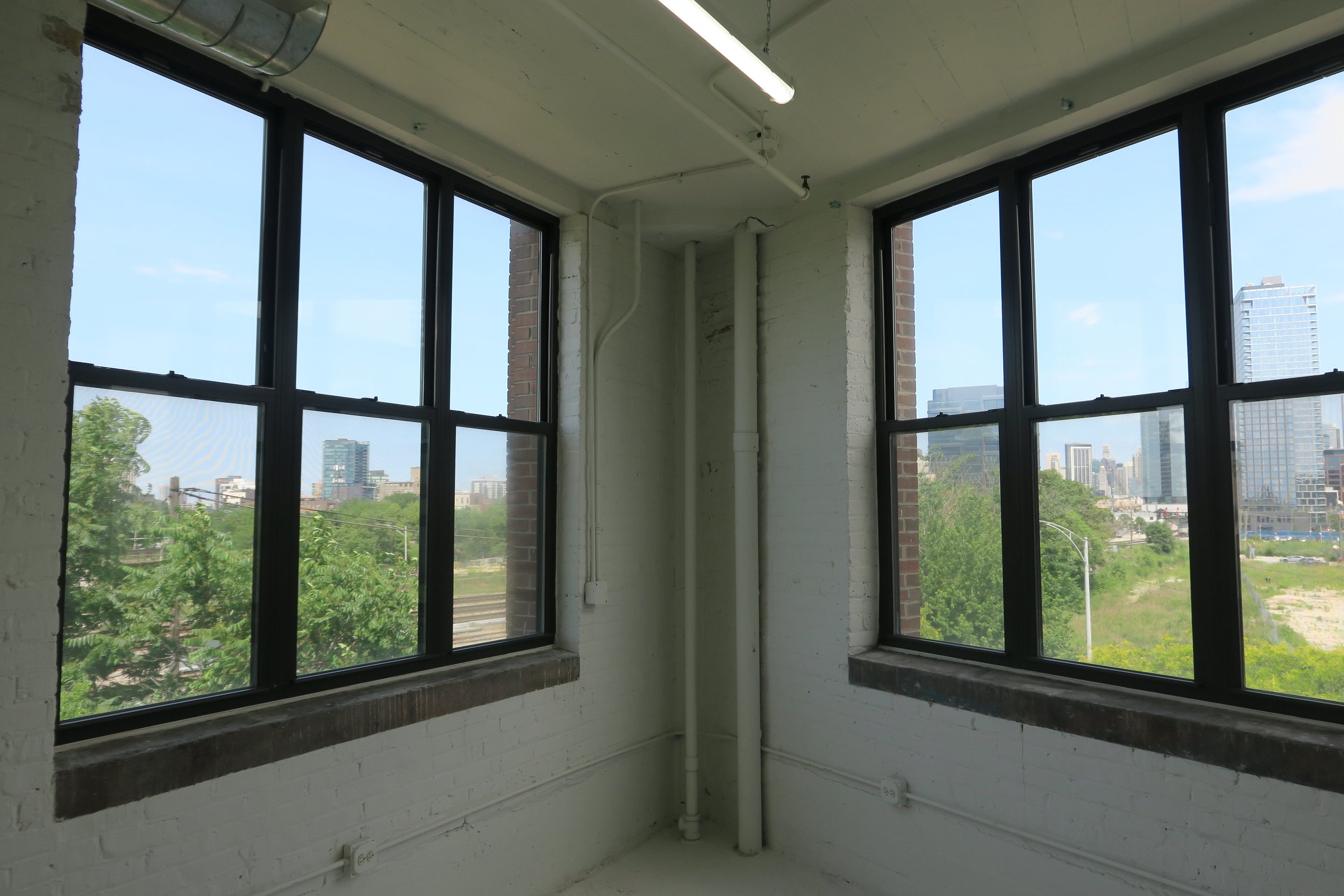 View of city skyline through two large industrial style windows in an unfinished room with white brick walls and visible electrical wiring.