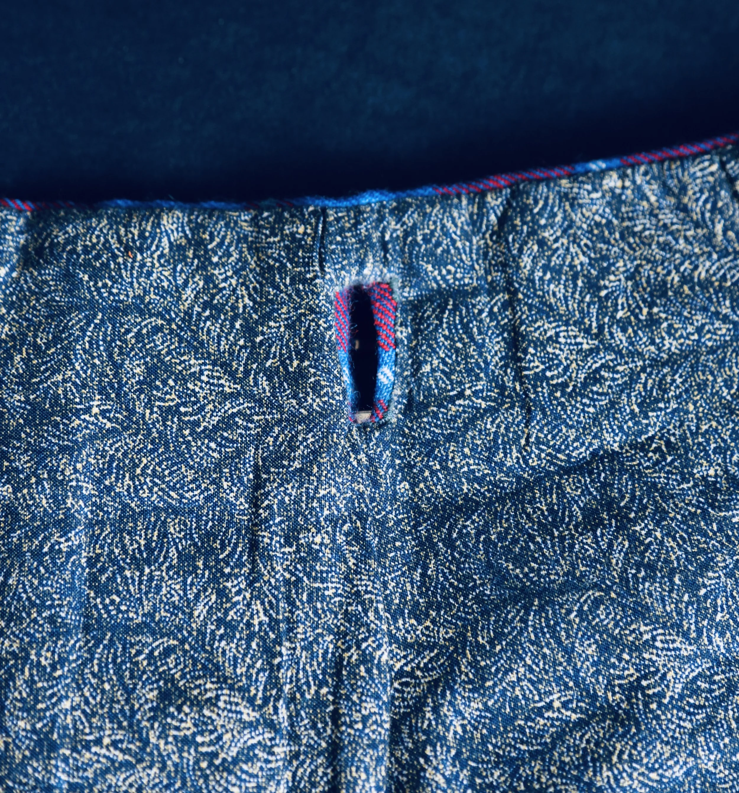 Close-up of the waistband of a pair of a handmade kilt with a buttonhole and red and blue striped stitching.