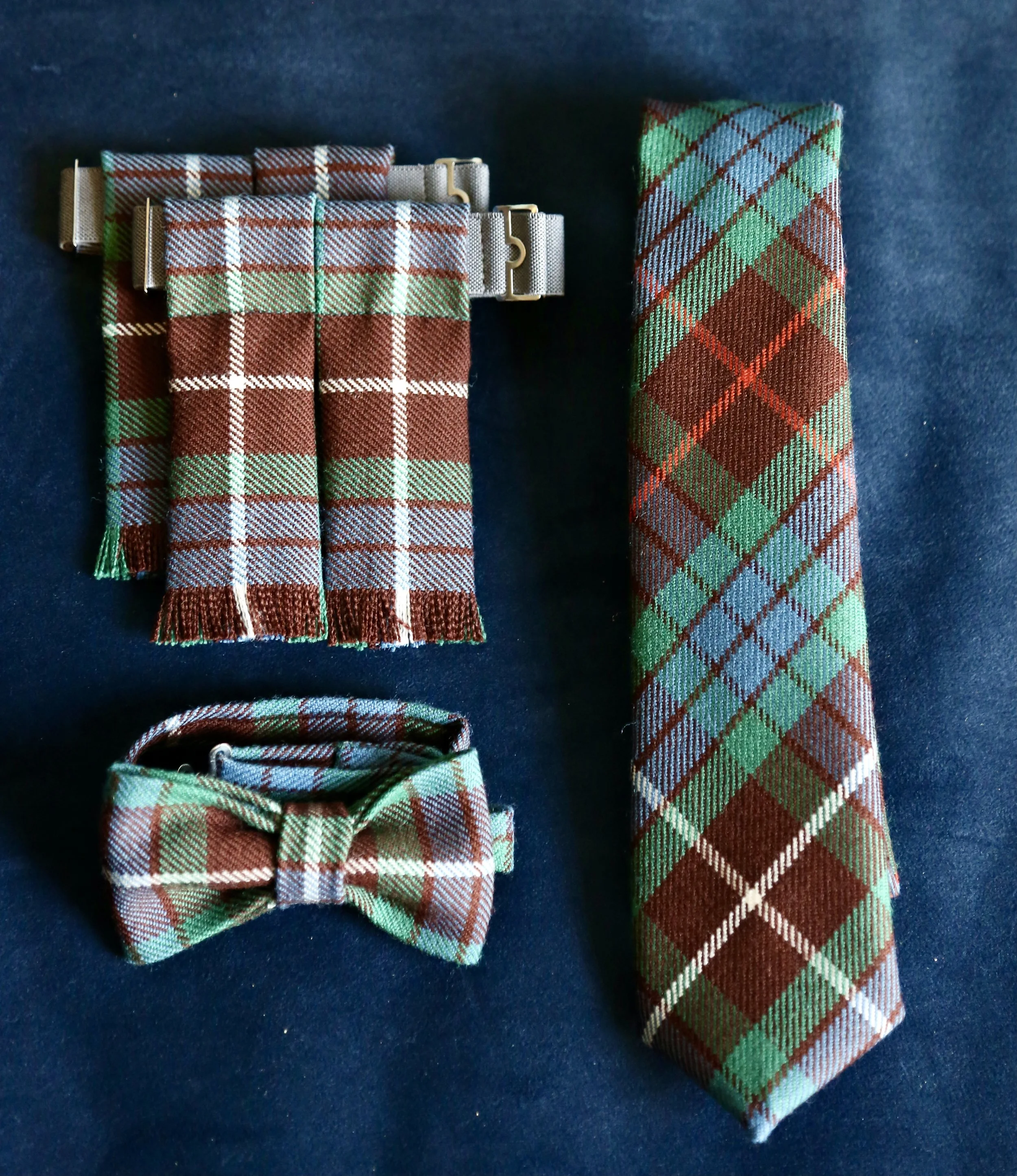 Set of tartan plaid accessories including a necktie, bow tie, and flashes, all with a green, red, blue, and white tartan pattern, laid on a dark surface.