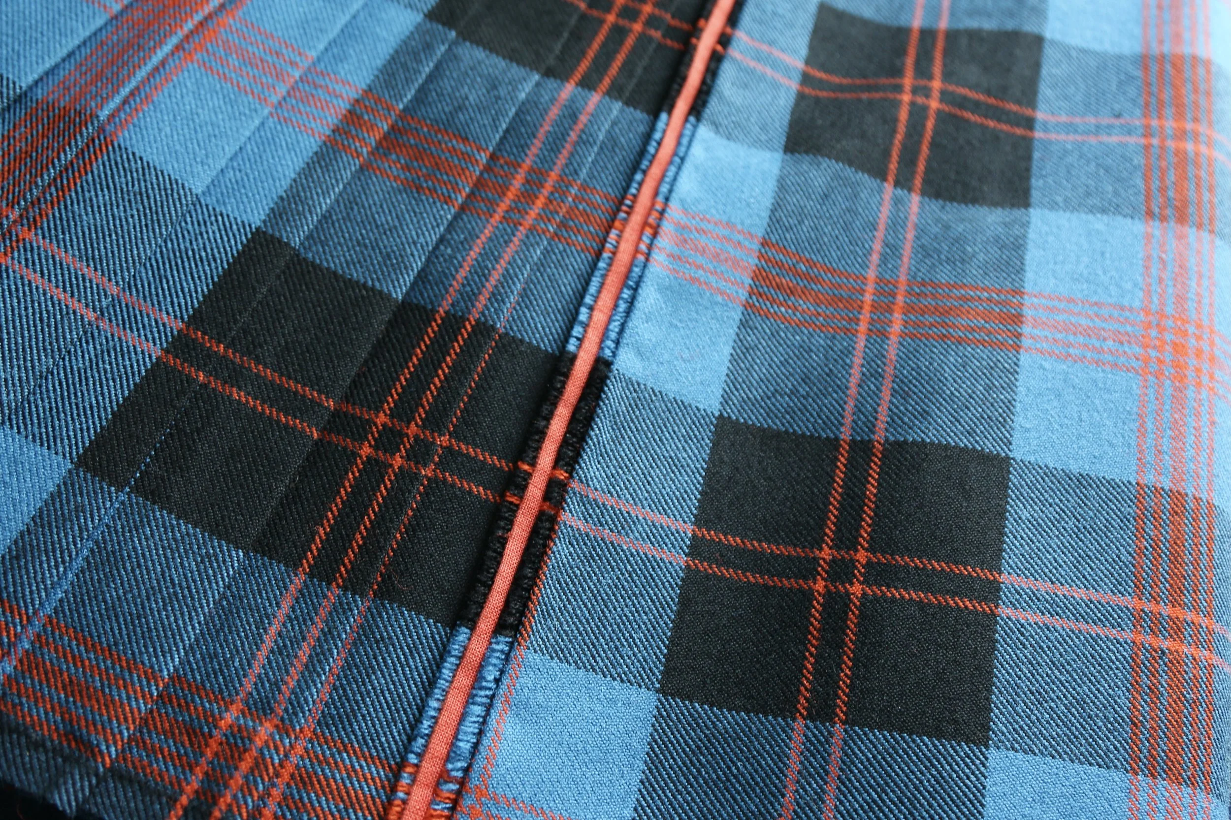 Close-up of a blue and black tartan patterned fabric with red lines, featuring a red orange accent in the center of the fringe.