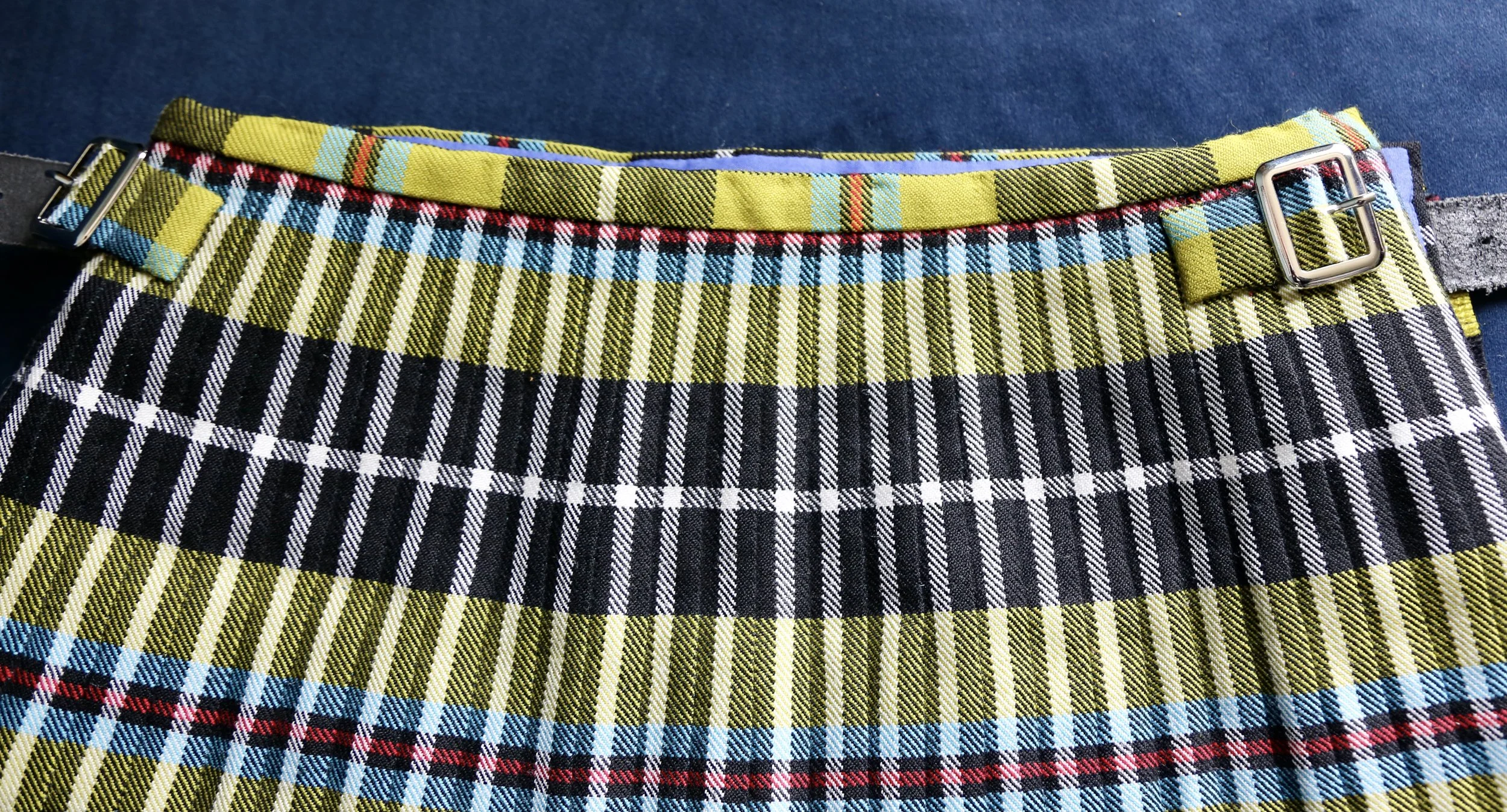 Close-up of a plaid/tartan kilt pleats with yellow, black, white, red, and blue pattern, shown with a black belt and buckle on a dark surface.