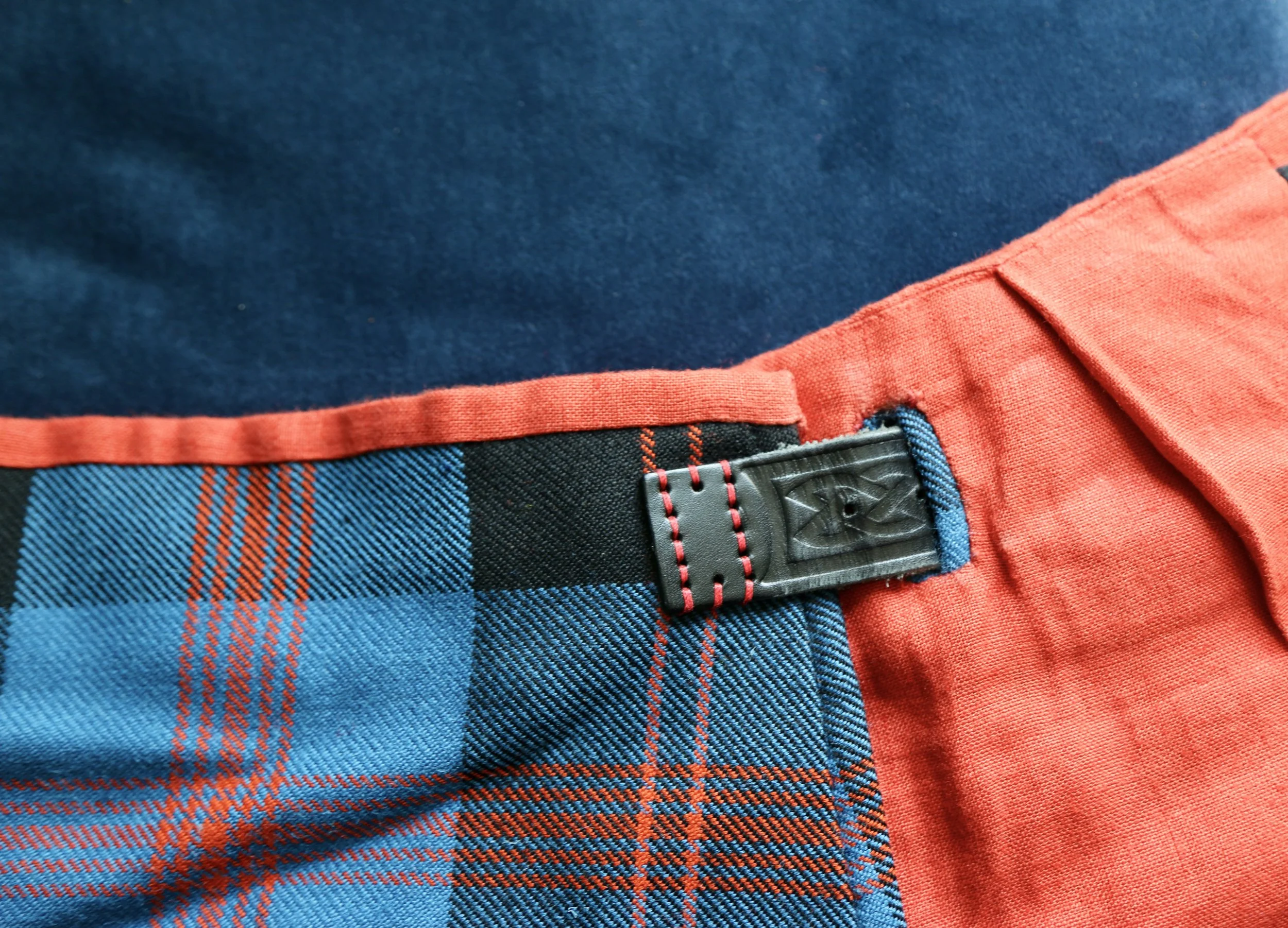 Close-up view of a colorful plaid fabric on clothing with a black adjustable belt with red stitching and a matte black strap, orange red lining  on a dark blue background.