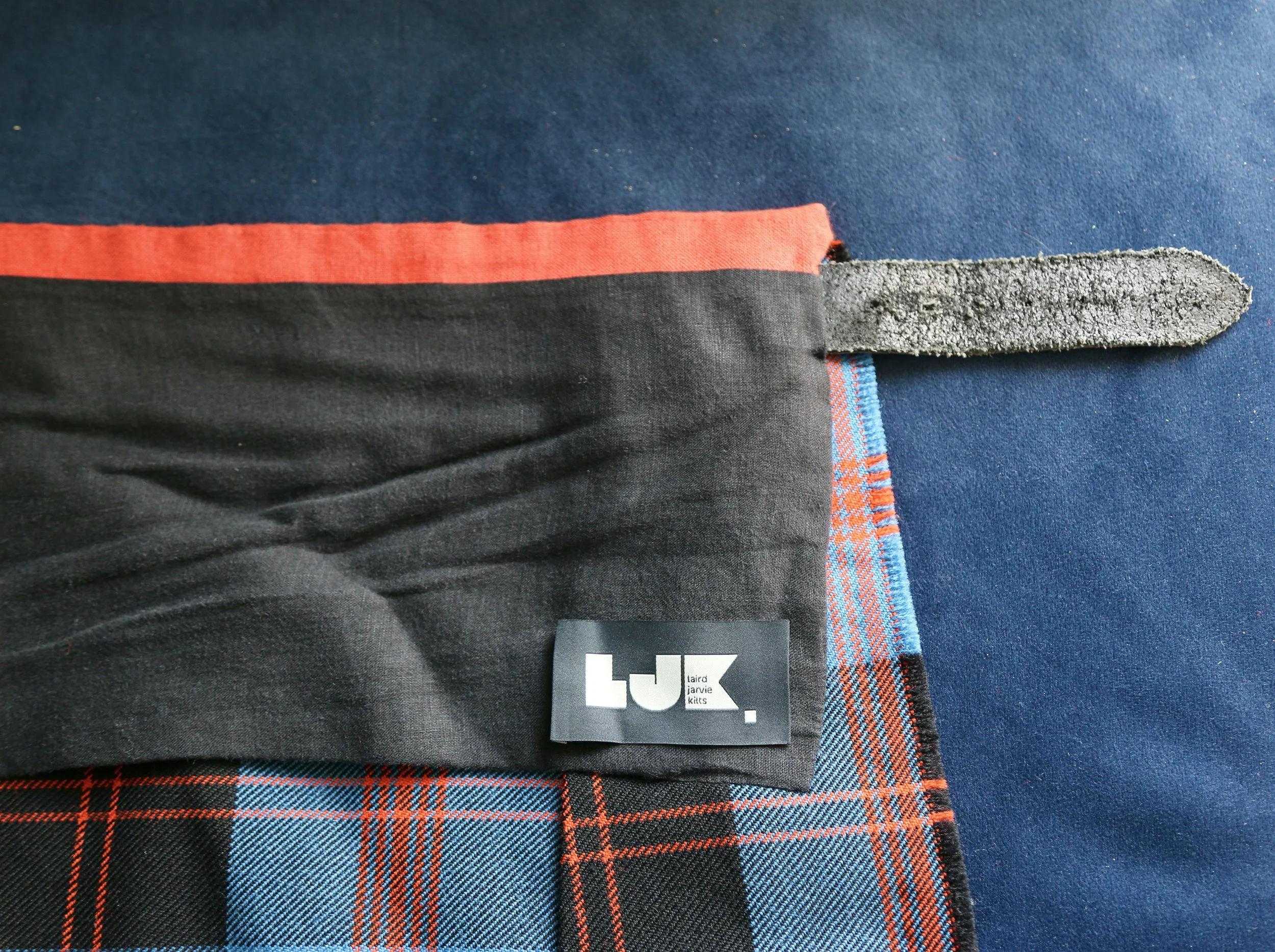 Close-up of a tartan plaid kilt lining with a black label reading 'LJ.K.' and a black leather strap, on a dark blue background.