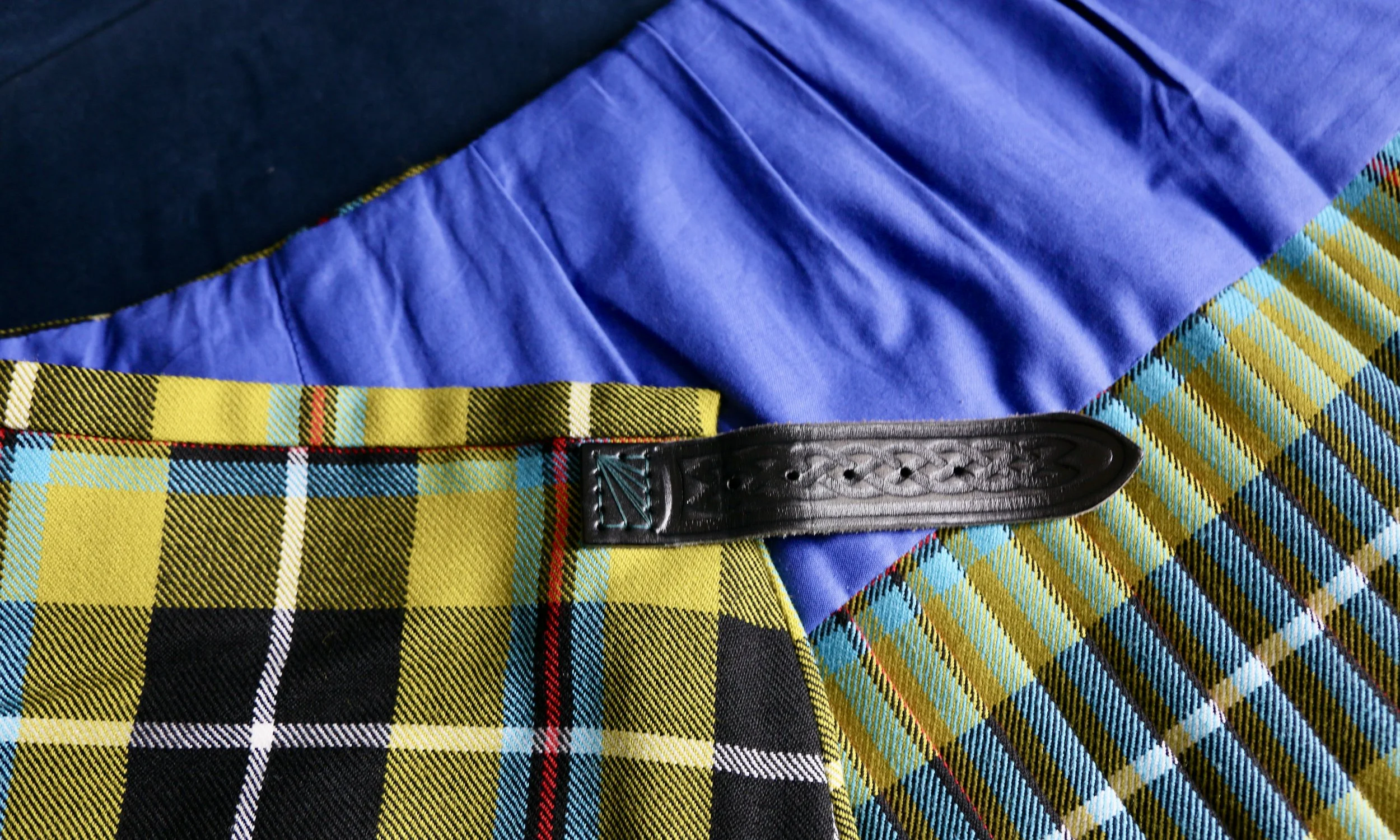 Close-up of a tartan/plaid kilt with a black leather strap and a blue fabric background.