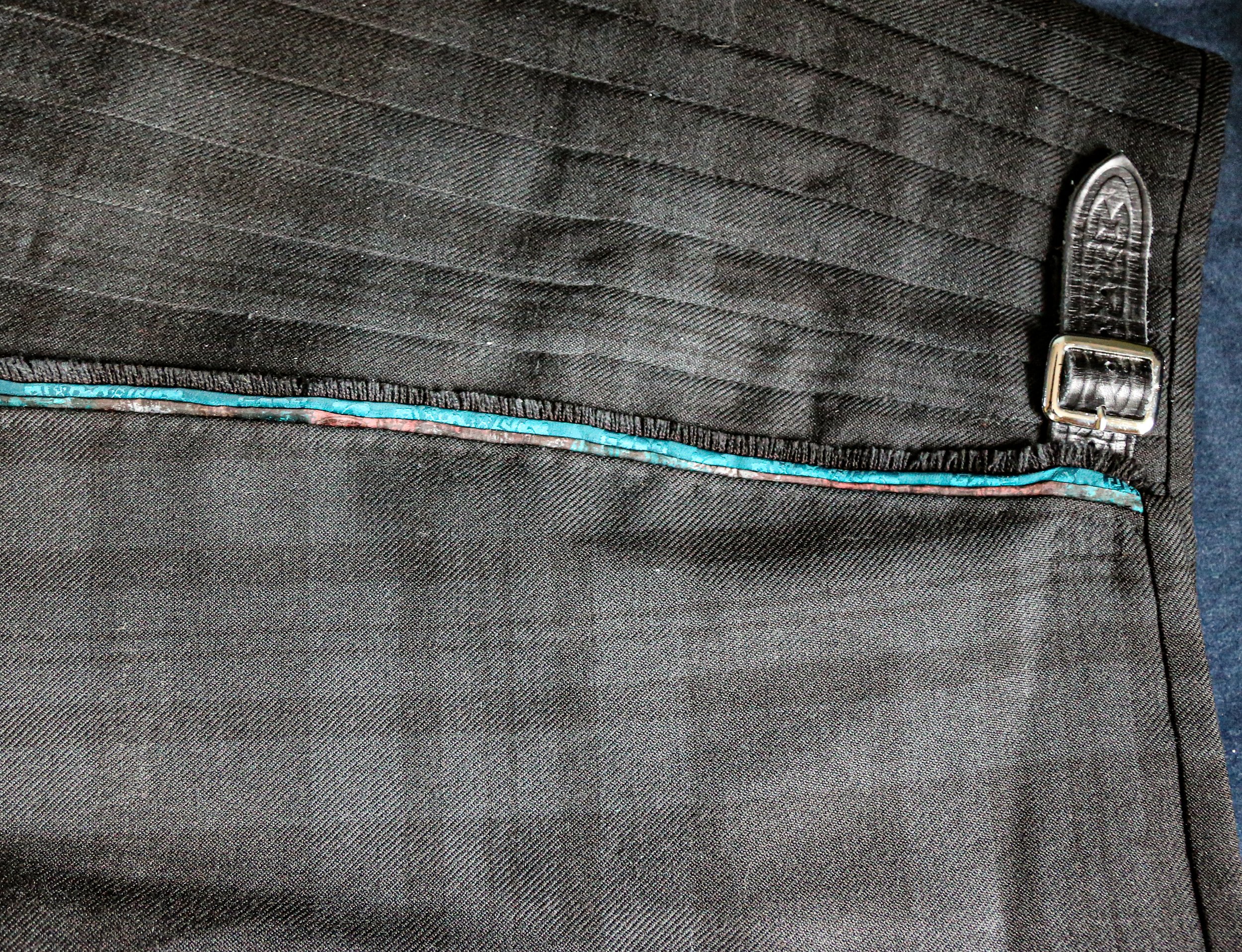 Close-up of a black fabric bag featuring a front pocket with a zipper that has a blue and pink trim, and a shiny silver hook on the corner.