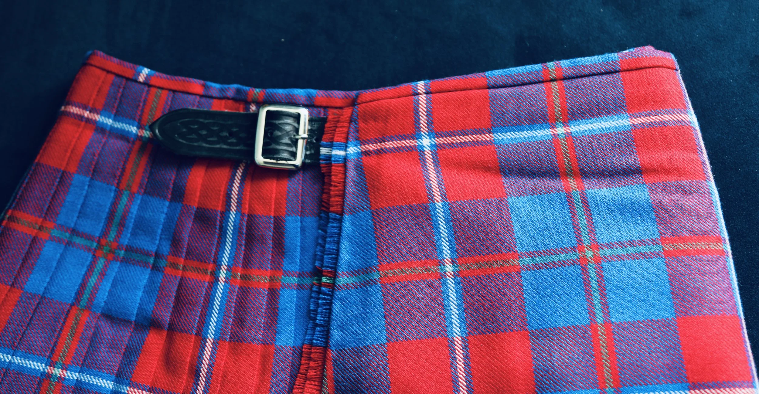 Close-up side view of red, blue, green, and white plaid fabric kilt with a black belt strap and silver buckle, on dark background.