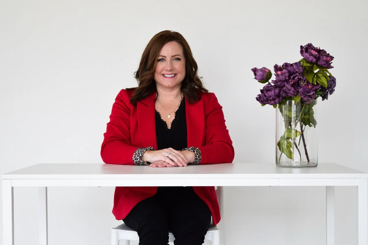 Strategic Psych contracted facilitator and licensed clinician, Olivia Moser, with brown hair, wearing a red blazer and black top, sitting at a white table with her hands folded. On the table, next to her, is a glass vase with purple flowers.