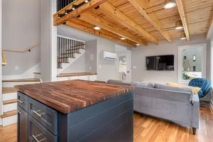 Open-concept living area with a wooden ceiling, gray sofas, a wall-mounted TV, and a kitchen island with a wooden countertop.
