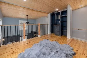 Empty room with wooden flooring, a blue bookshelf, a gray wall, a ceiling fan, and a gray furry rug.