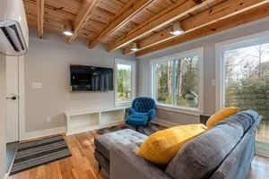 Living room with large windows, gray sofa with yellow pillows, blue armchair, TV mounted on the wall, wooden ceiling, hardwood floor, and white built-in shelf.