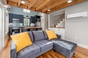 Living room with a gray sectional sofa with yellow pillows, wooden ceiling, and a kitchen area in the background.