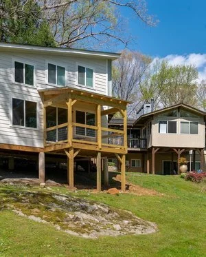 View of a house with a wooden deck on stilts, overlooking a grassy yard, with neighboring house in the background and partly cloudy sky.