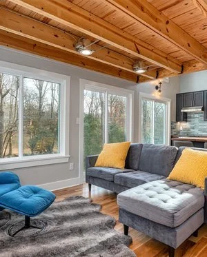 Living room with large windows, gray sofa with yellow pillows, blue lounge chair, and wooden ceiling beams.