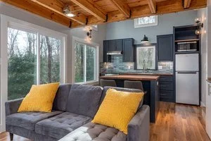 Living room with large windows, gray sofa with yellow pillows, wooden ceiling, and small kitchen area with dark cabinetry and stainless steel appliances.