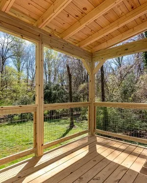 View of a screened-in porch with wooden flooring and frame, overlooking a wooded backyard with trees and grass under a sunny sky.