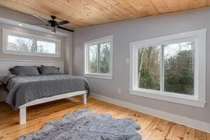 A bedroom with a bed, two large windows, and a wooden ceiling, featuring a gray rug on a wooden floor.