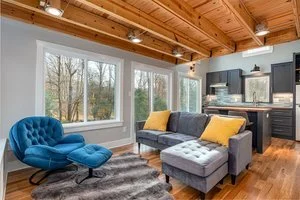 Living room with large windows, blue chair, gray and yellow sofa, wooden ceiling, and kitchen in the background