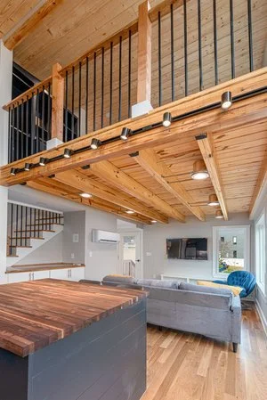 Living room with wooden ceiling, exposed beams, and stairs leading to a second-floor balcony. There's a gray sectional sofa, a wall-mounted TV, and a blue accent chair. Hardwood floors and large windows let in natural light.