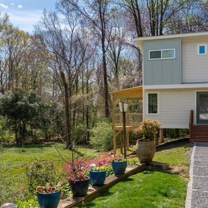 A backyard garden with potted plants on a wooden edge, a grassy lawn, and a house with a small porch and stairs, surrounded by trees.