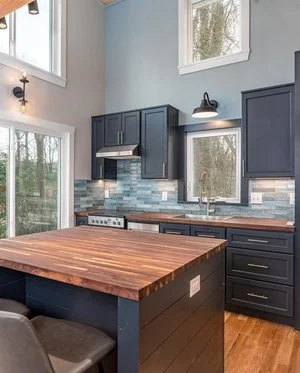 Kitchen with dark blue cabinets, a wooden island, and a blue tiled backsplash. There are two windows letting in natural light and a stovetop with oven. A sink is visible under one window.