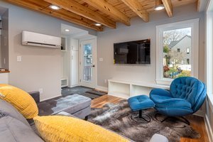 Living room with wooden ceiling beams, white walls, a wall-mounted TV, a window, a glass door, a yellow pillow on a sofa, a blue lounge chair, and a gray and white area rug.