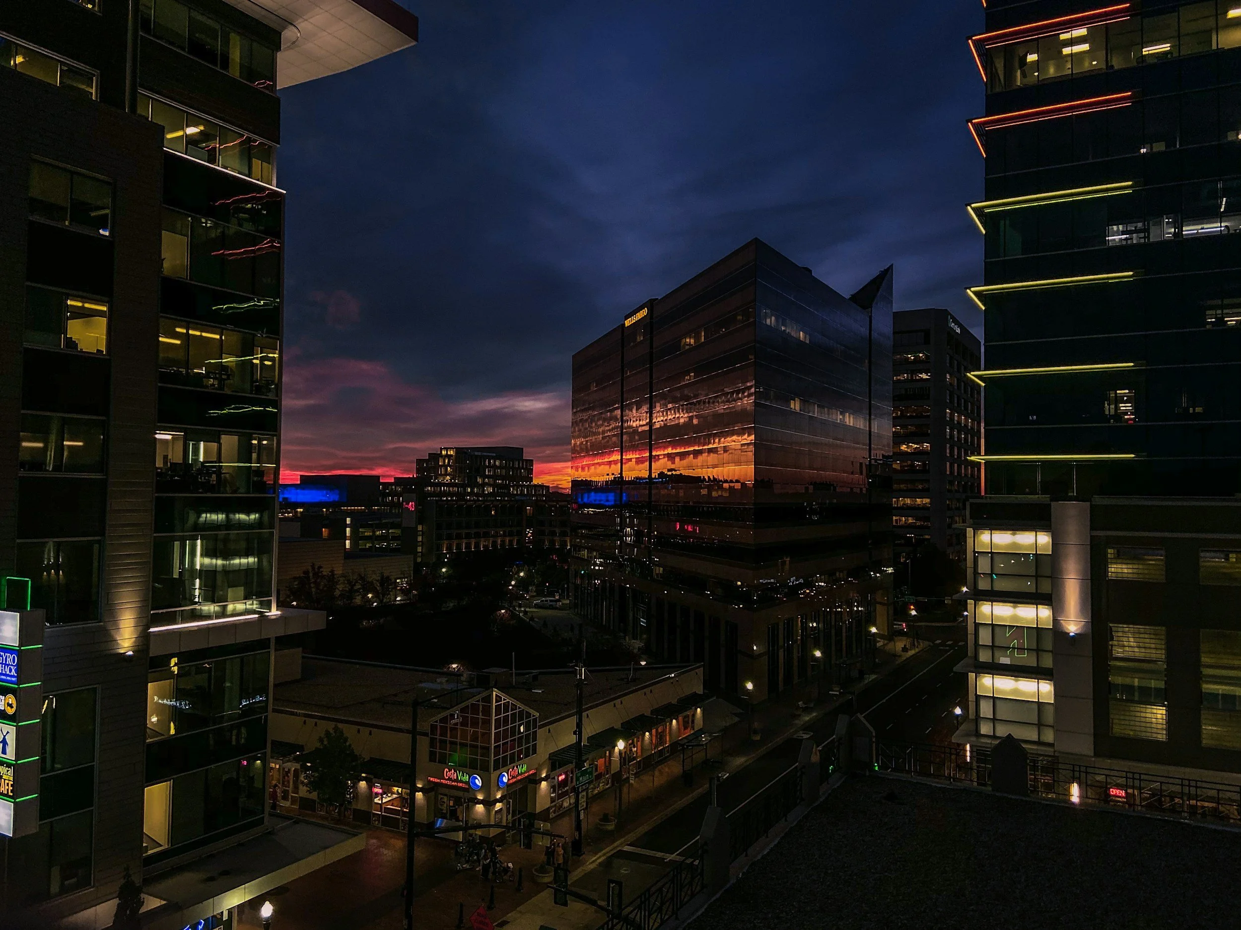Nighttime view of modern office buildings with illuminated outlines and glass windows, a colorful sunset sky, and a busy shopping area below.