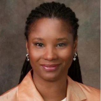 A woman with dark hair styled in braids, wearing pearl earrings and a beige top, smiling at the camera.