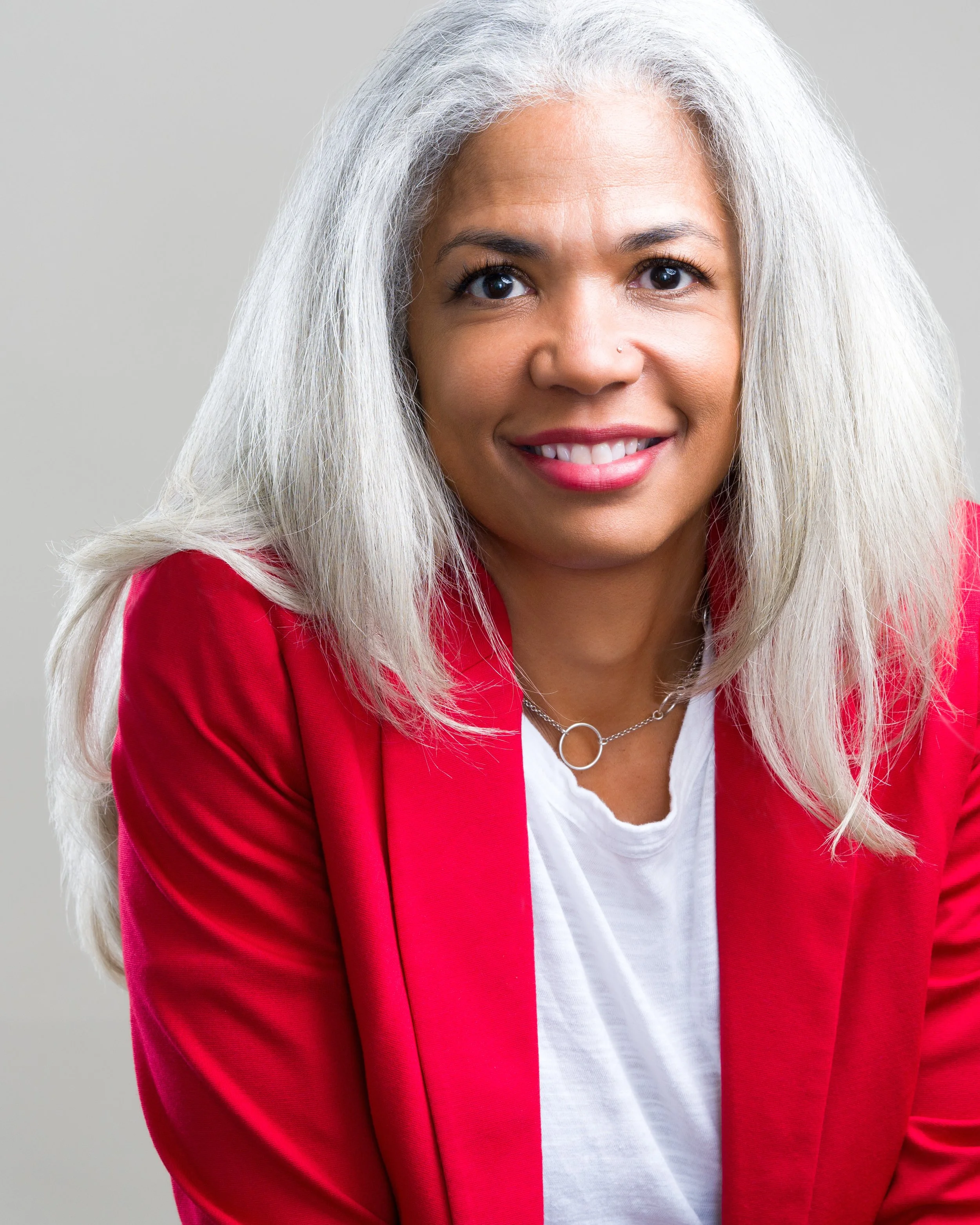 A woman with long, gray hair, wearing a red blazer and white shirt, smiling at the camera against a plain background.